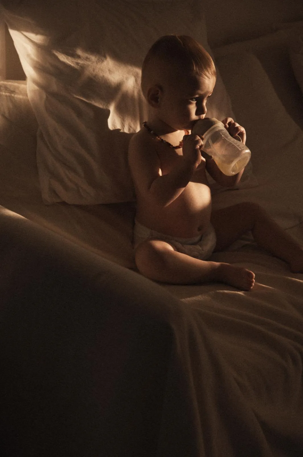 A baby sitting up on a bed with a cup, lit by a single window, everything else fallen away to shadow