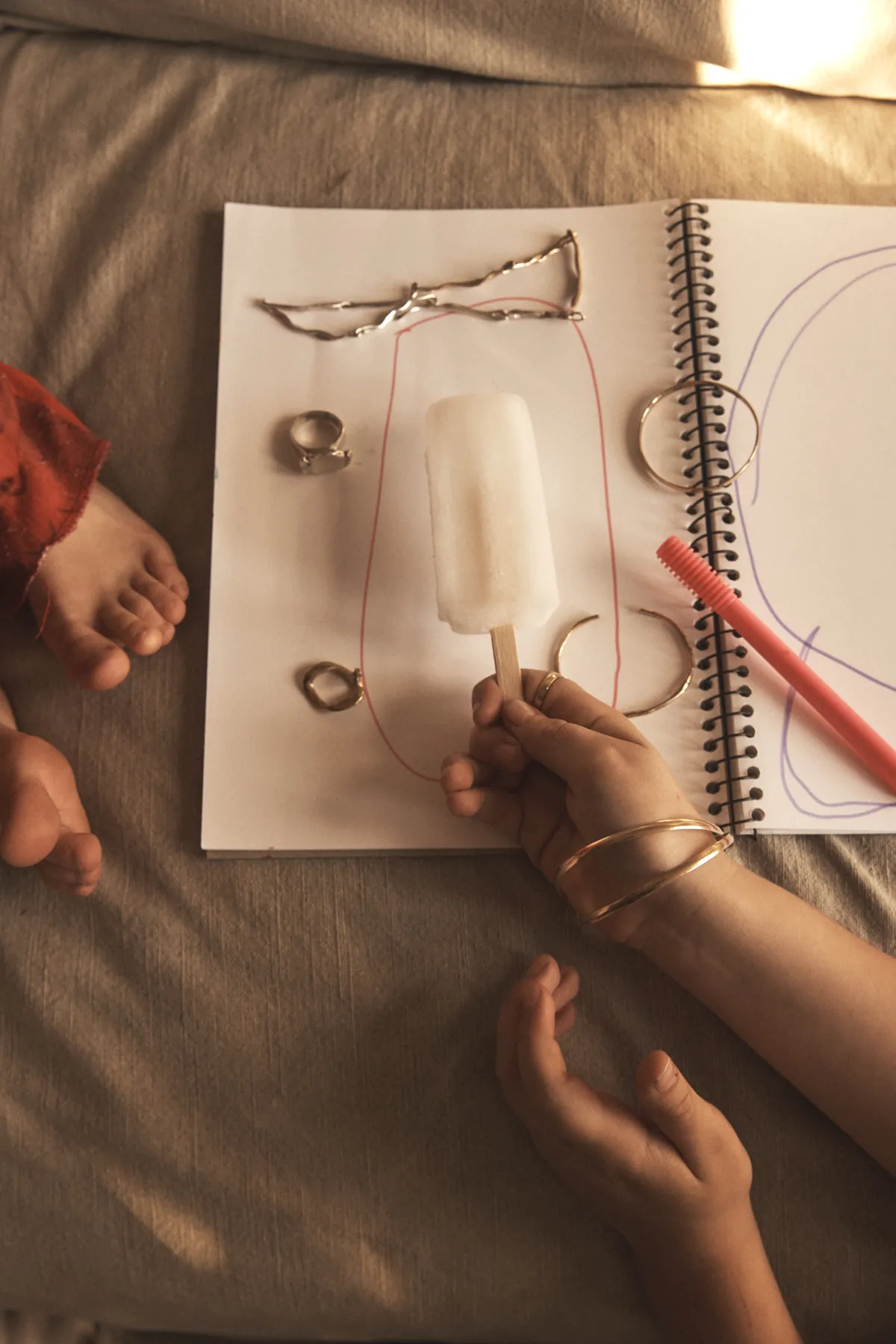 Seen from above: an ice block on an open sketchbook, scribbled drawings, rings laid out, small bare feet at the edge of the frame