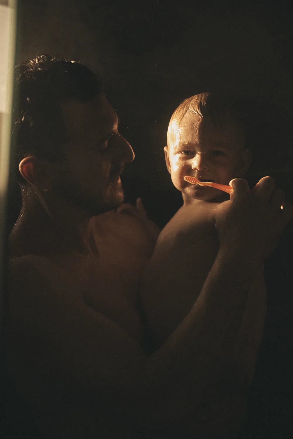 A father holding his toddler in a dark bathroom, brushing the child’s teeth, one strip of warm light across their faces