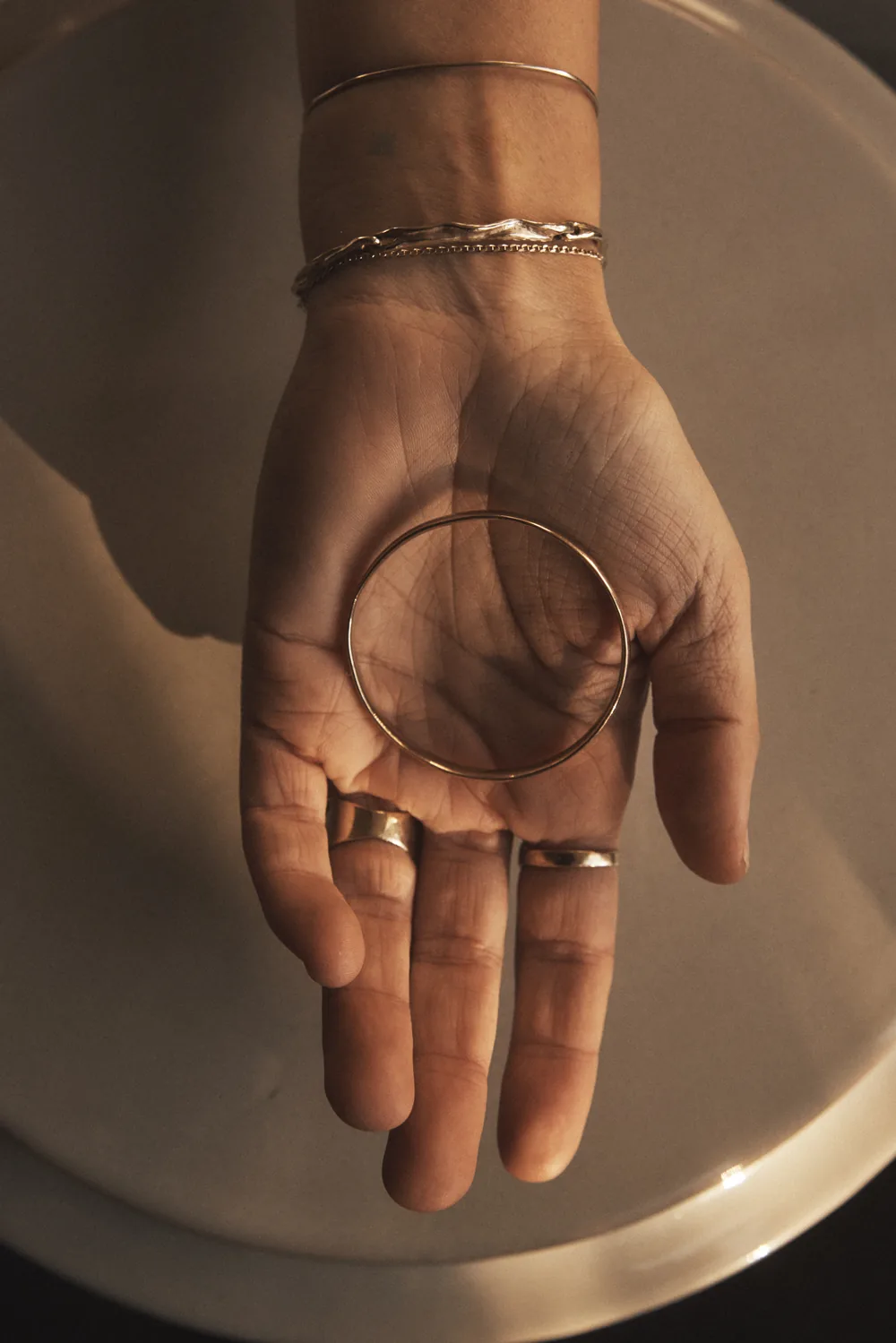 An open palm resting in a bowl, a fine bangle lying across it, rings on two fingers, late sun raking across the skin