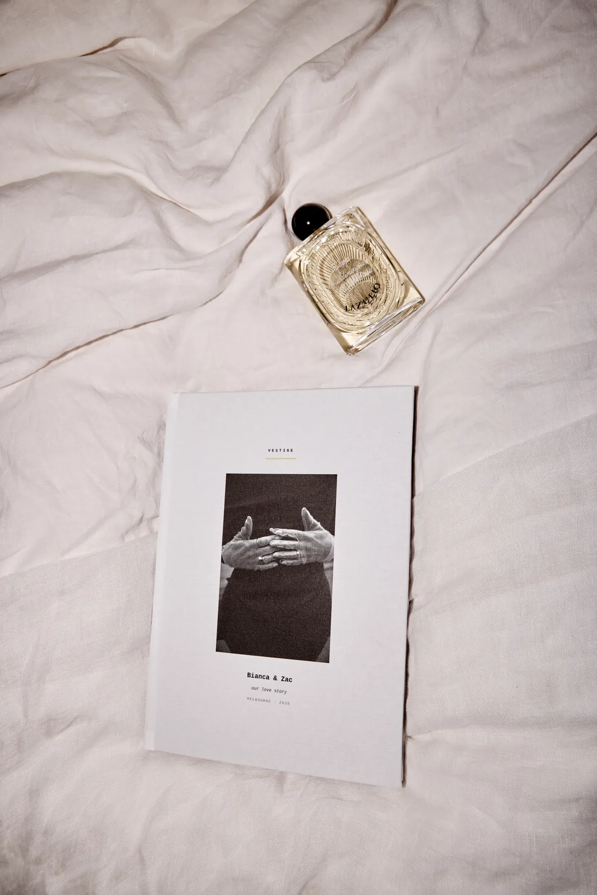 A Vestige book laid on linen bedding beside a bottle of perfume