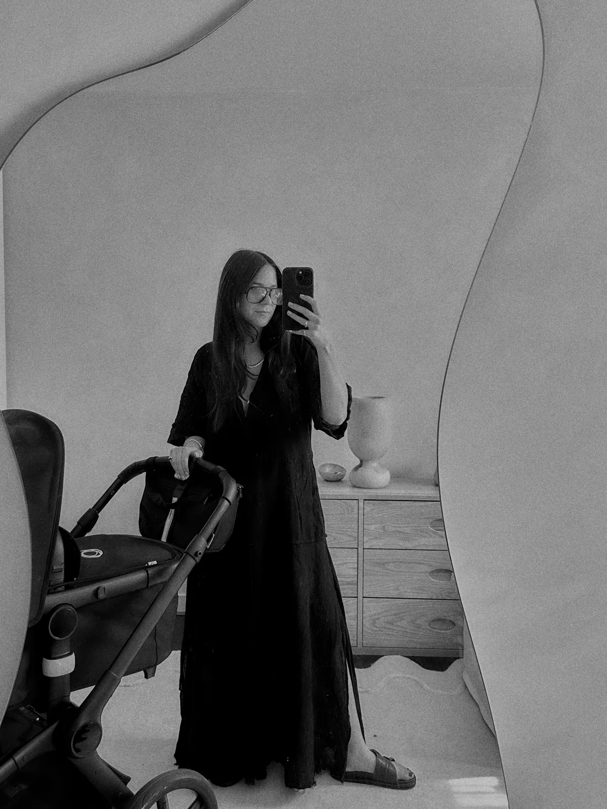 Liane Hurvitz photographing herself in a mirror at home, pram beside her — black and white